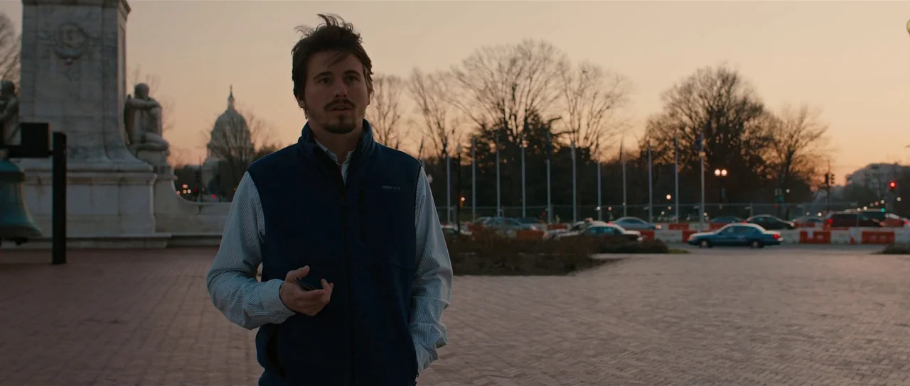 The East (2013) movie still: medium — A brick-paved plaza sits before a government building dome in the late afternoon. A man in a blue vest walks…