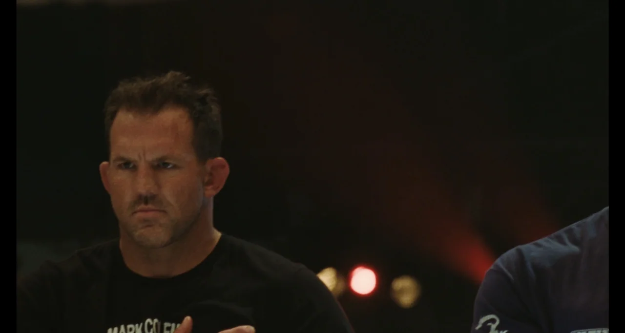 The Smashing Machine (2025) movie still: medium — An dimly lit arena interior with dark shadows concealing the background. A man in a black shirt…
