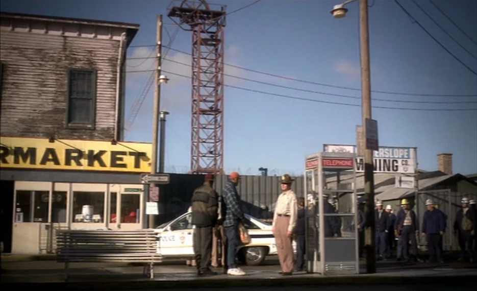 Dear Wendy (2005) movie still: wide — An industrial town square features a weathered building with a prominent yellow supermarket sign. A group of…