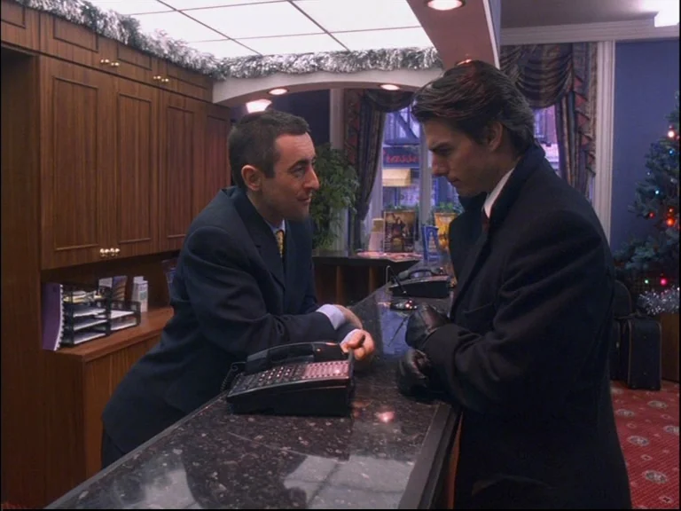 Eyes Wide Shut (1999) movie still: medium — A hotel check-in counter featuring wood-paneled walls and a granite countertop. A man in a suit speaks…