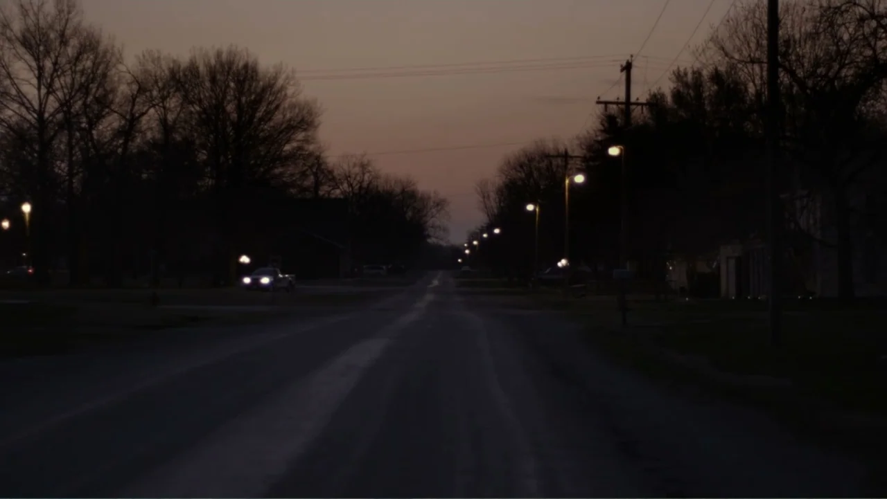 Rich Hill (2014) movie still: establishing — A quiet residential street is lined with mature trees and illuminated by distant streetlamps. A car…