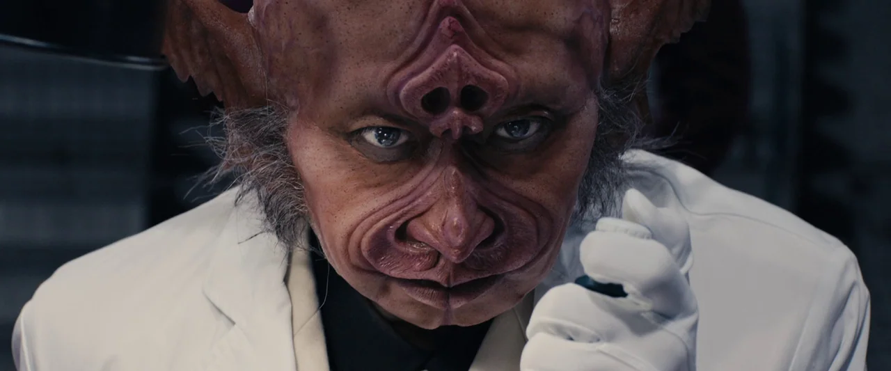 Shin Kamen Rider (2023) movie still: closeup — A close-up shot of an alien character wearing a white lab coat. The character stares directly into…