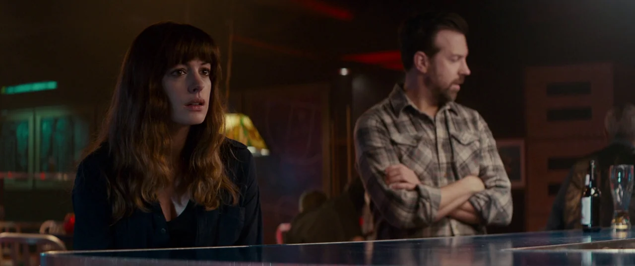 Colossal (2016) movie still: medium — A dimly lit neighborhood bar features dark wood surfaces and scattered warm practical lighting. A man and a…