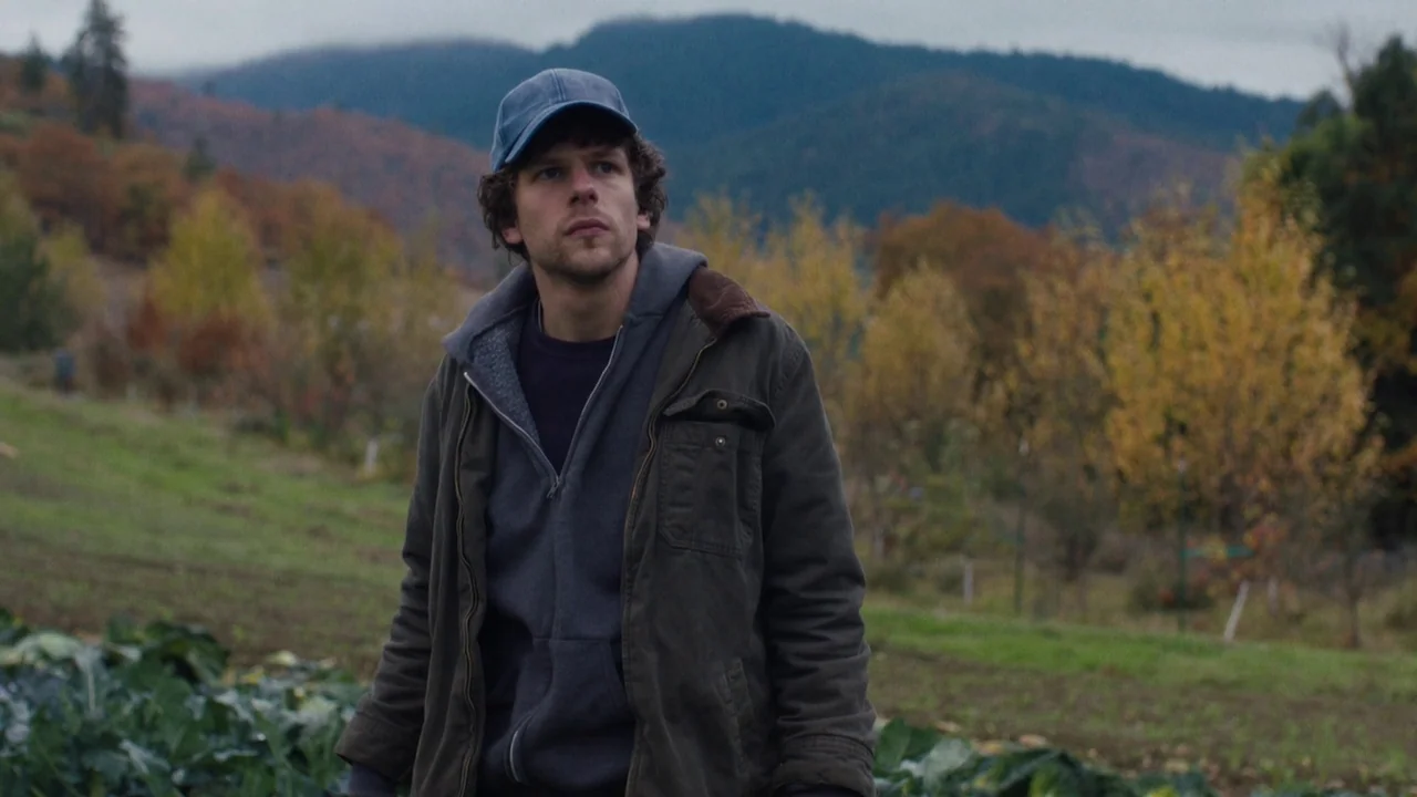 Night Moves (2013) movie still: medium — An open field at the base of rolling hills under a grey cloudy sky. A man stands in the foreground wearing…