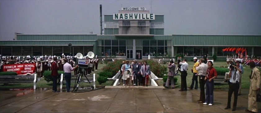 Nashville (1975) movie still: wide — A 1970s airport terminal features a large, prominent 'Welcome to Nashville' sign on roof. A group of people…