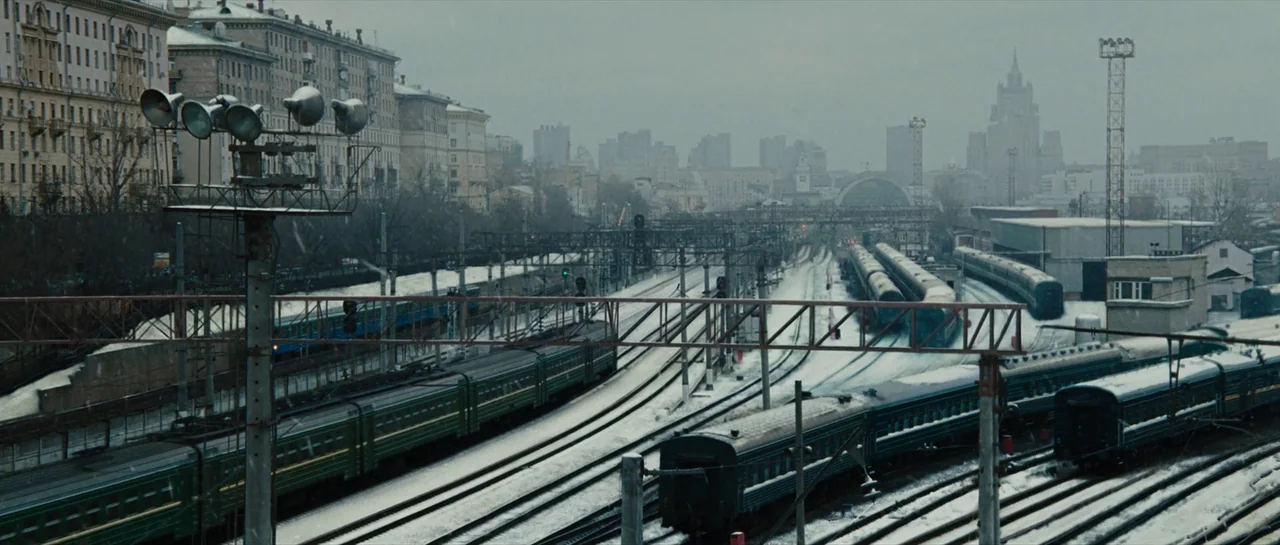 Iron Man 2 (2010) movie still: establishing — An expansive railway yard filled with parked passenger trains sits under a cloudy sky. Light snow…