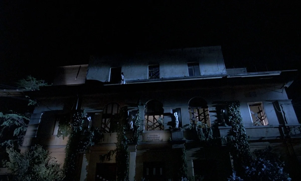 Phenomena (1985) movie still: wide — An old, dilapidated stone building is covered in dense vines and boarded-up windows at night. The structure is…
