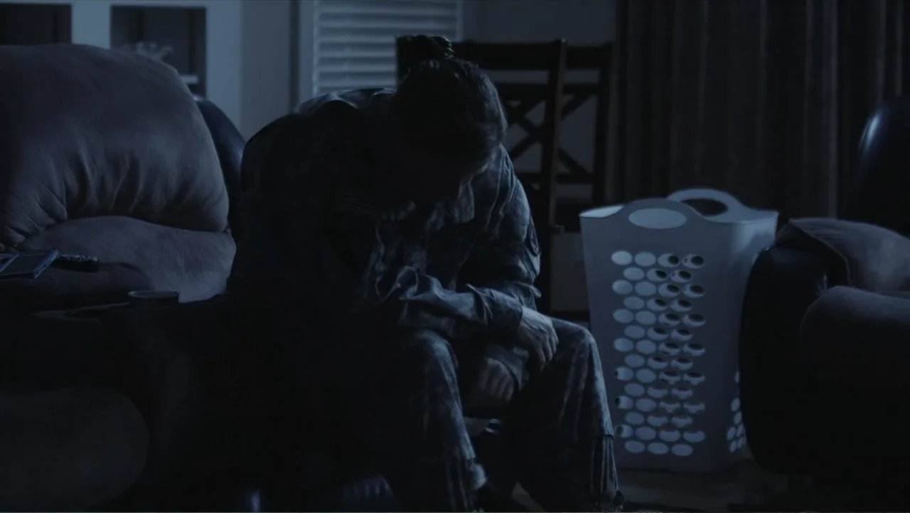Death of a Child (2017) movie still: medium — A dimly lit living room at night contains a couch and a plastic laundry basket. A person wearing a…