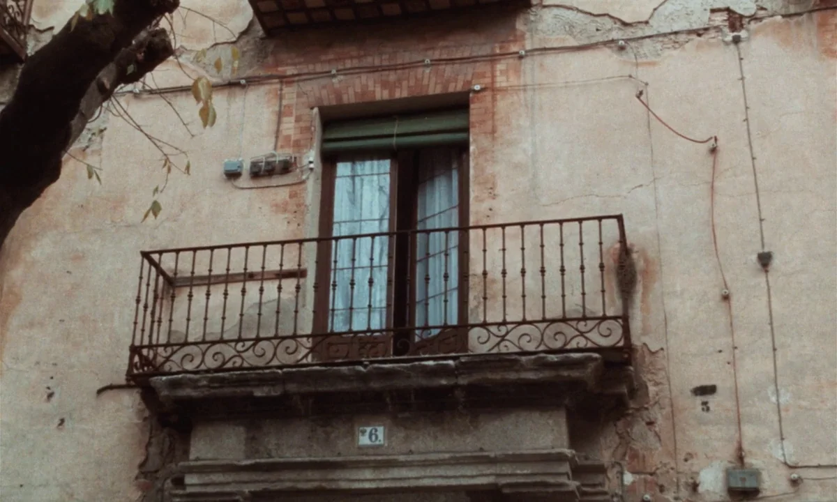 Tristana (1970) movie still: medium — A weathered stone building exterior features a metal balcony with an open window. The faded stucco wall is…