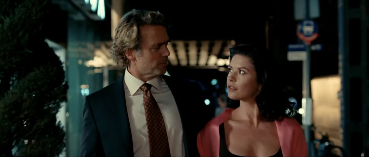 The Rebound (2009) movie still: medium — A dark city street at night with a blurred building facade in the background. A man in a suit and a woman…