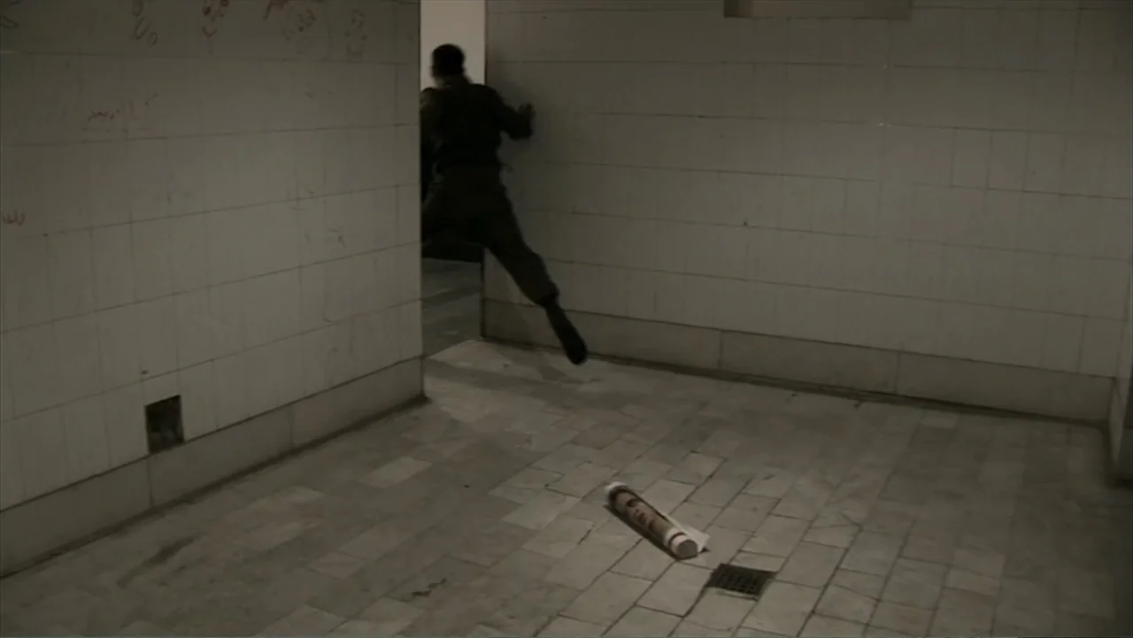 Offside (2015) movie still: wide — A stark, tile-walled public restroom features graffiti on the walls and a floor drain. A lone figure moves…