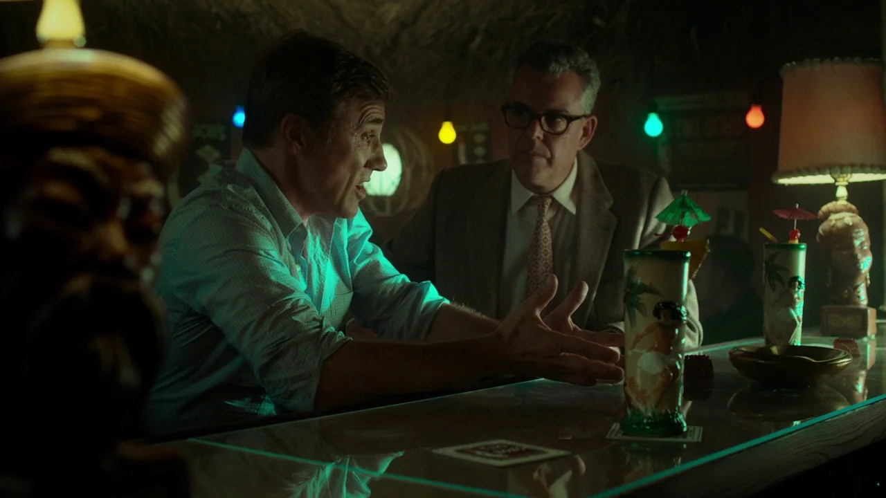 Big Eyes (2014) movie still: medium — A dimly lit tiki bar features thatched roofing and decorative colored lighting bulbs overhead. Two men sit at…