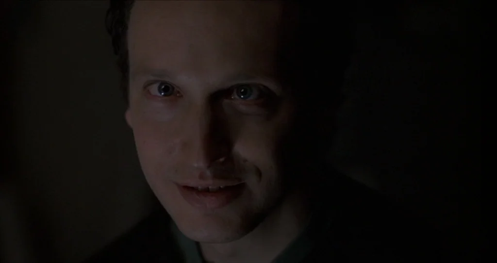 The Silence Of The Lambs (1991) movie still: closeup — A man is centered in a dimly lit, undefined interior space. He gazes directly into the lens…