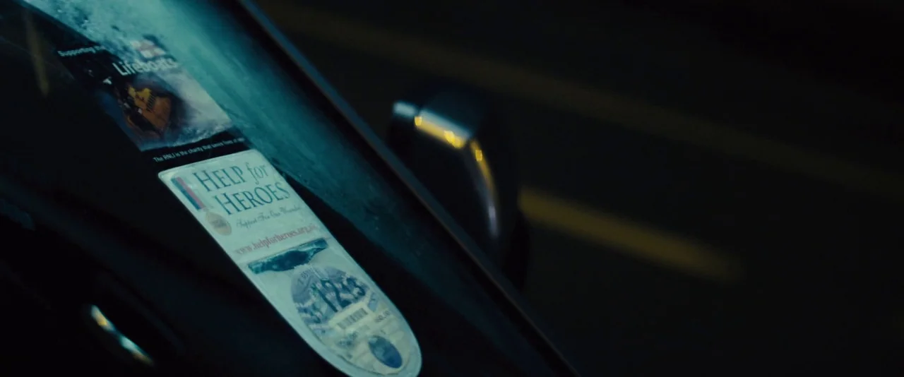 Locke (2013) movie still: closeup — A close view of a dark car windshield at night displays charity stickers. The glass surface reflects ambient…