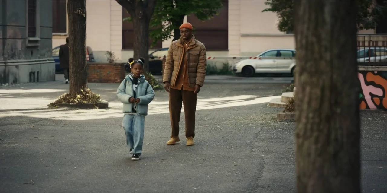 Autumn Beat (2022) movie still: fullbody — An asphalt clearing between buildings with several trees and a distant car. An adult man walks alongside…