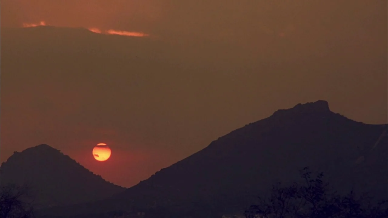 The Rapture (1991) movie still: establishing — A dark silhouette of jagged mountains rises against a hazy, dusk-colored sky. A bright red sun dips…