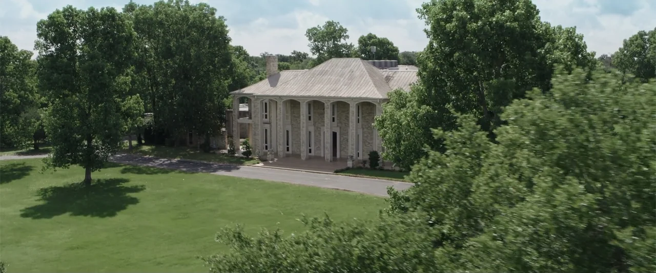 Playing God (2021) movie still: establishing — A two-story stone mansion with a metal roof sits among lush green trees. A paved driveway leads to…