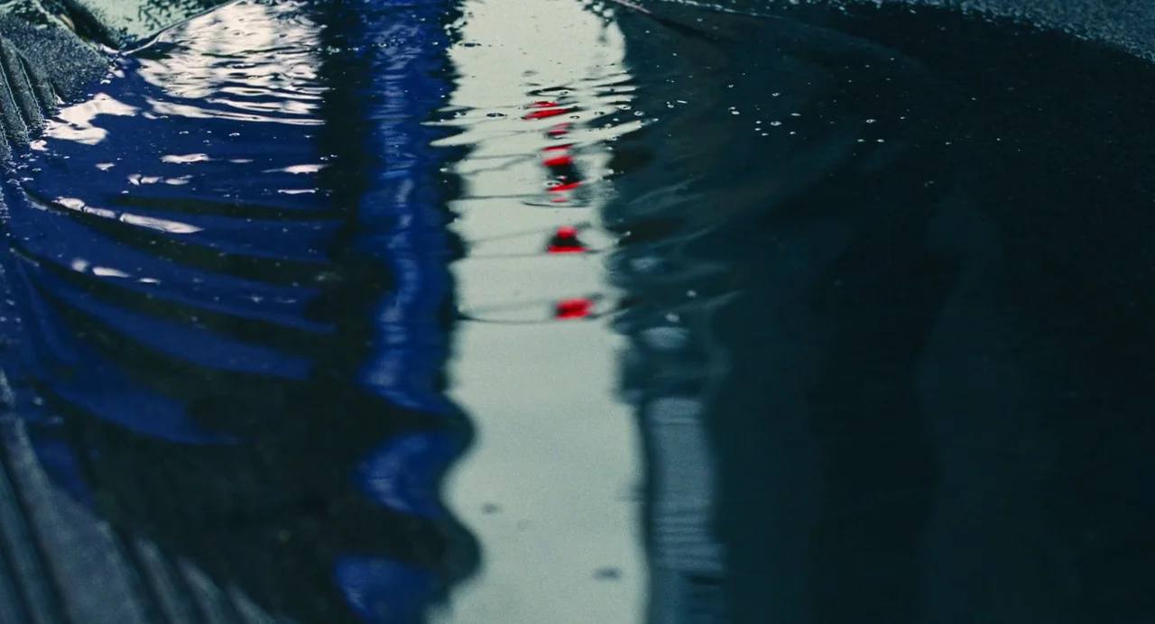 Past Lives (2023) movie still: closeup — A dark puddle on asphalt captures blurry reflections of surrounding urban lights. A line of small, red…