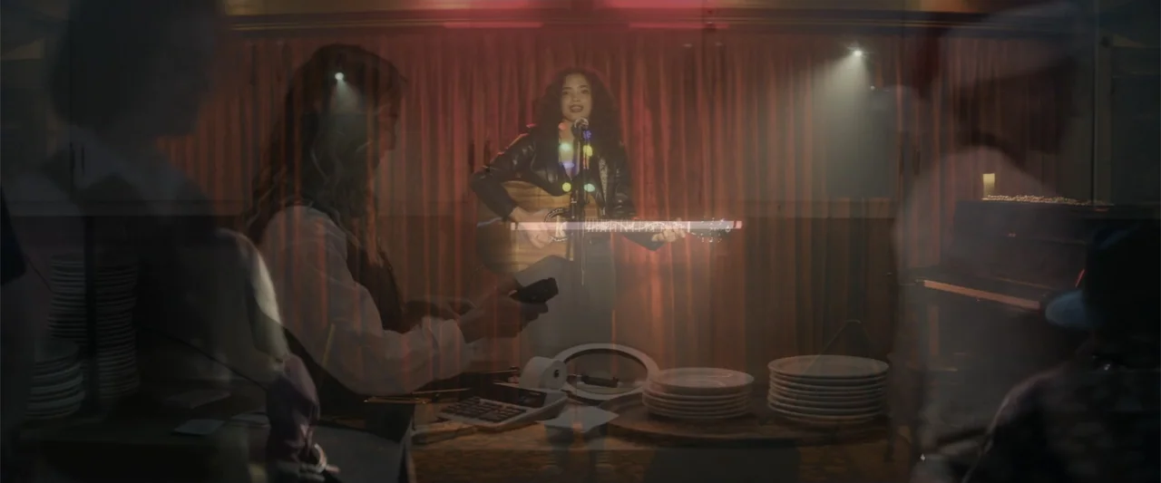 Holiday Harmony (2022) movie still: medium — A dimly lit dining area features a performer on stage against a dark curtain backdrop. A person works…