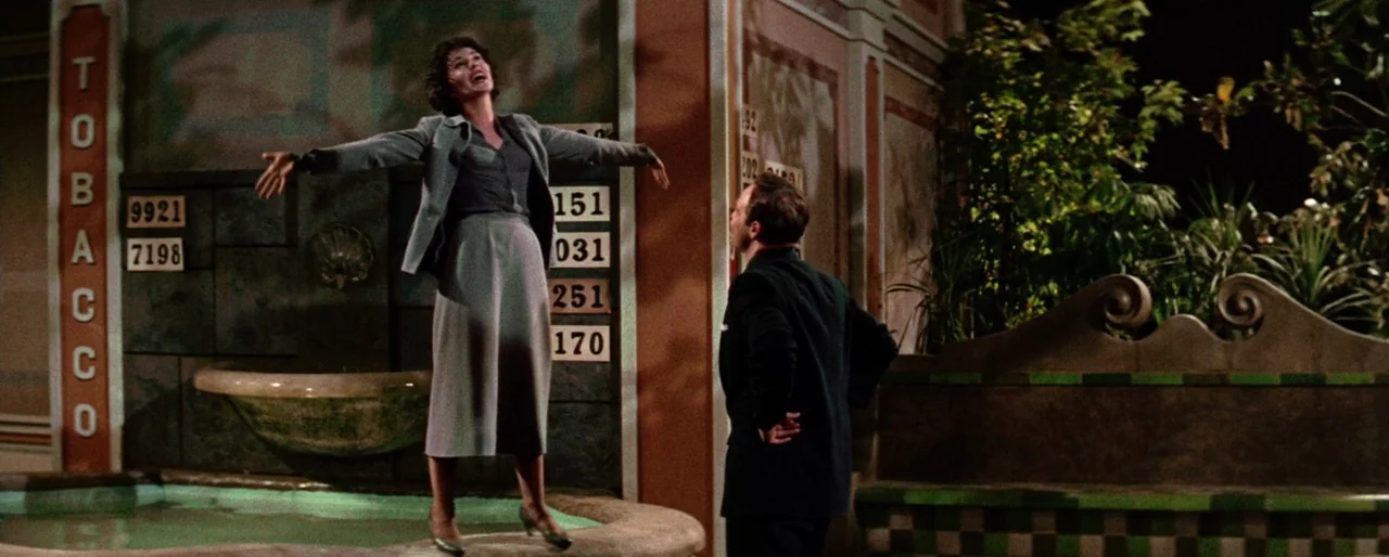 Guys and Dolls (1955) movie still: fullbody — A nighttime outdoor courtyard features a fountain with tiled walls and numbered placards. A woman…