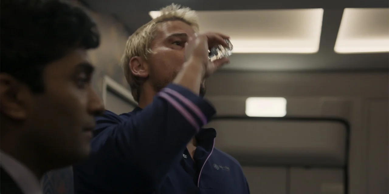 Fight or Flight (2025) movie still: medium — A dimly lit indoor passageway with plain wall panels and recessed ceiling lights. A blonde man drinks…