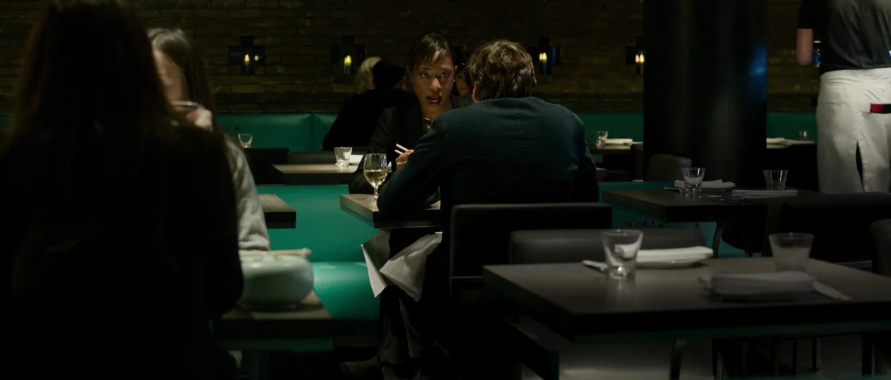 The Face of an Angel (2014) movie still: medium — A dimly lit restaurant features dark brick walls and teal leather booths. A woman and man sit at a…