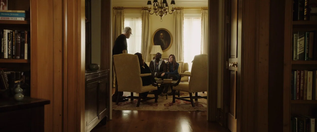 Get Out (2017) movie still: wide — A view through a wooden doorway into a sunlit library filled with bookshelves. Four individuals interact while…