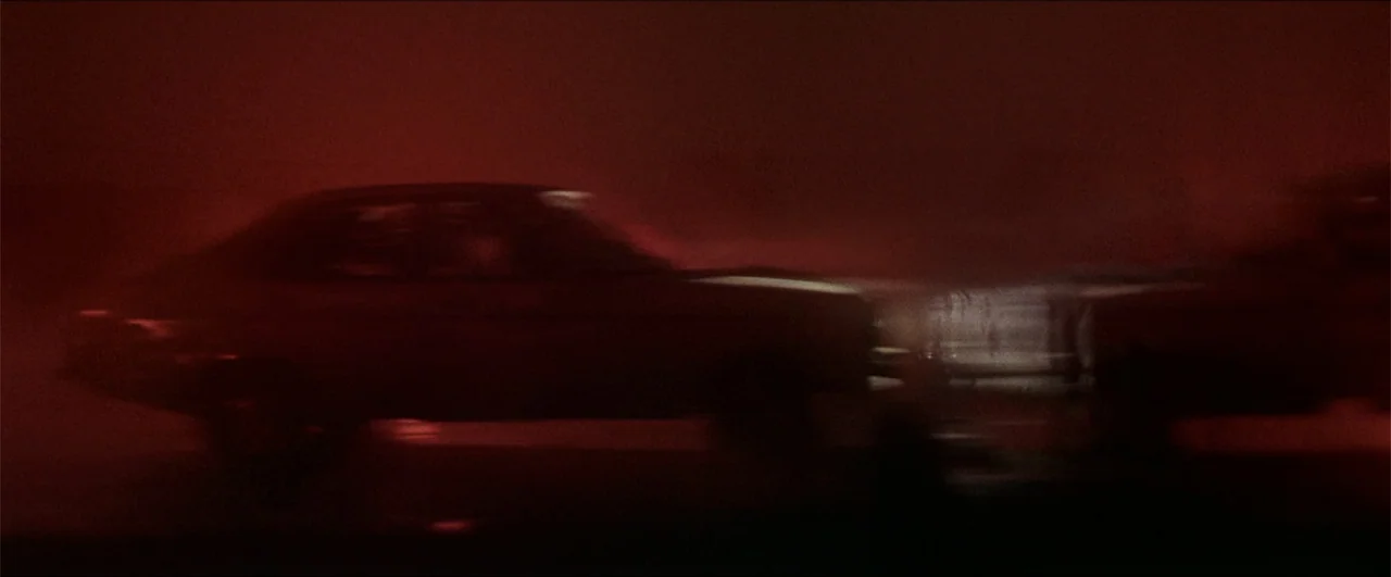 Better Man (2024) movie still: medium — A dark road at night is engulfed in a thick red atmospheric light. A blurred vehicle speeds through the…