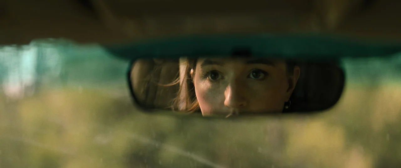 No One Will Save You (2023) movie still: closeup — A close-up view through a vehicle's rearview mirror shows a young woman's reflection. The…