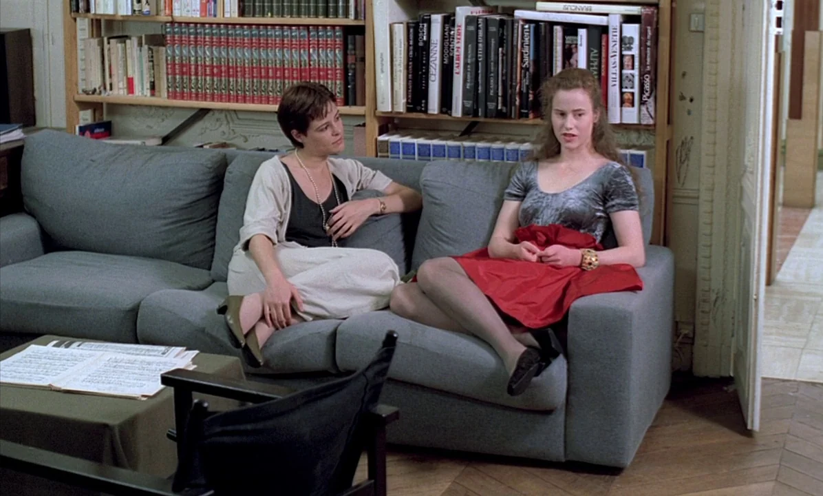 A Tale of Springtime (1990) movie still: medium — A room with filled wooden bookshelves and a grey sofa occupies the space. Two women sit on the…