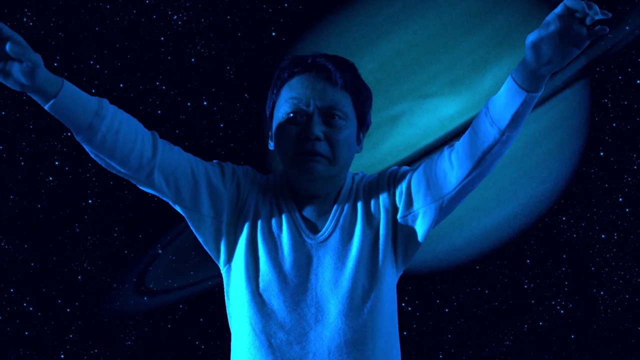The Happiness of the Katakuris (2001) movie still: medium — A person floats against a backdrop of a large ringed planet and starry sky. The man has…