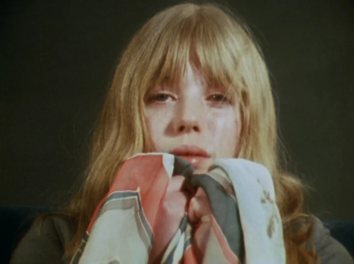 Lucifer Rising (1972) movie still: closeup — A person is centered in a dimly lit, dark space with no visible environment. She holds a patterned…