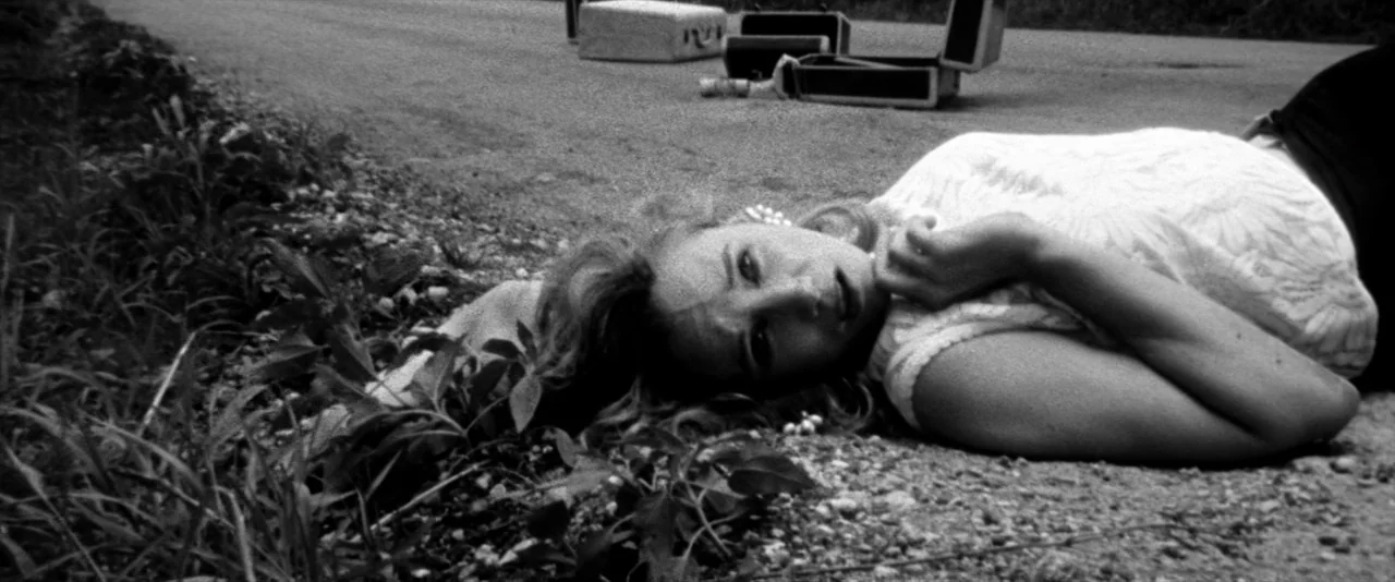 JFK (1991) movie still: medium — A gravel road shoulder bordered by grass with open suitcases in the distance. A woman lies on the ground in a white…