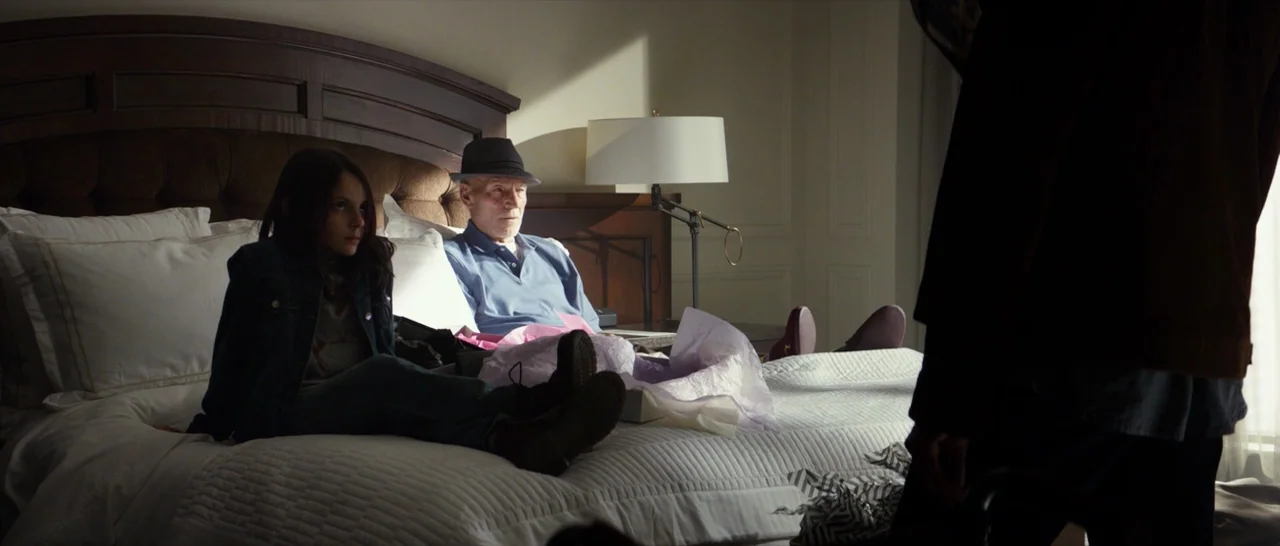 Logan (2017) movie still: medium — A hotel room features a large bed with white linens and a dark wooden headboard. An elderly man and a young girl…