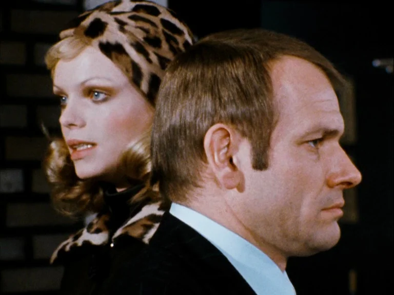 Making of: World On A Wire (1973) movie still: closeup — Two individuals stand in profile against a dark background in an interior space. The woman…