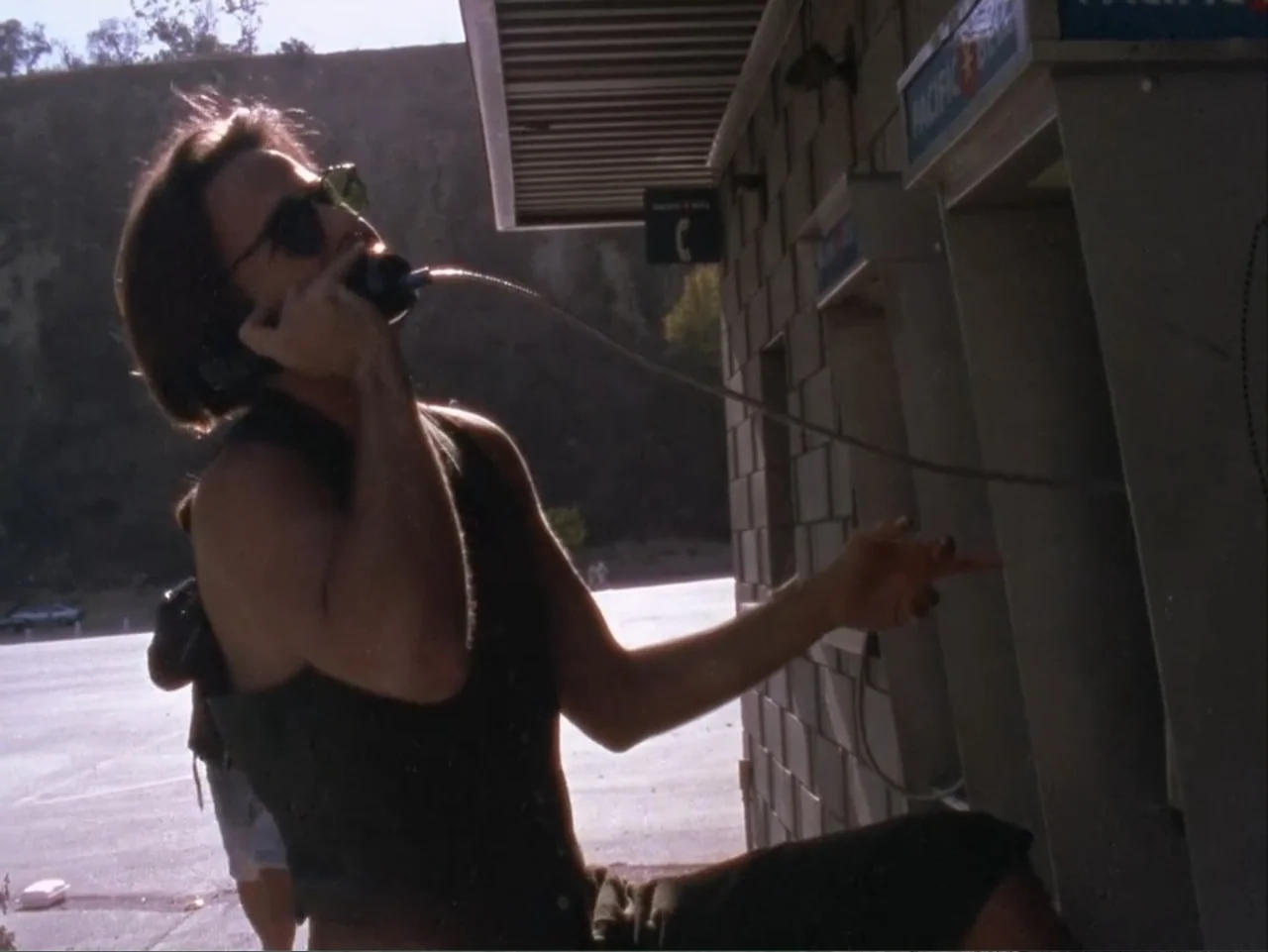 The Living End (1992) movie still: medium — An outdoor concrete structure with a wall-mounted payphone stands in bright daylight. A person holds the…