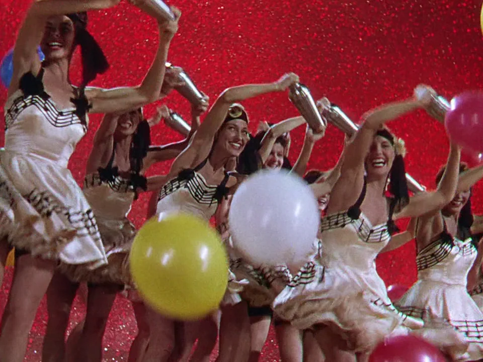 Singin’ in the Rain (1952) movie still: medium — A group of dancers performs on a brightly lit stage with a red background. The women wear matching…