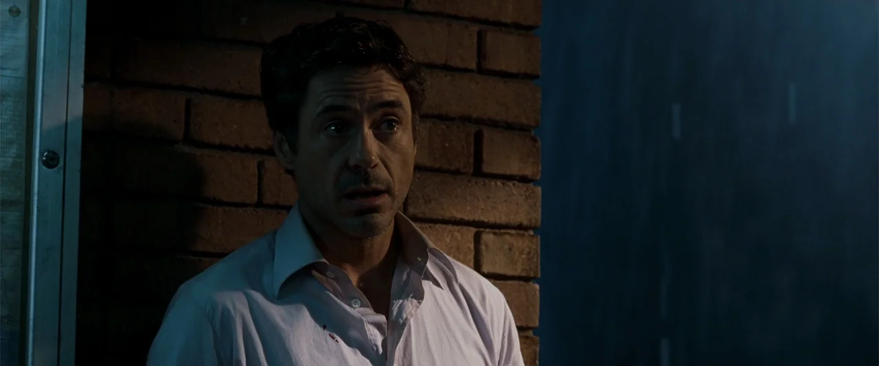 Due Date (2010) movie still: medium — A man stands in a dimly lit corridor against a red brick wall. The subject wears a white dress shirt with a…
