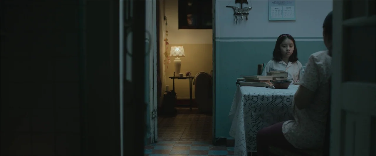 Radical (2023) movie still: medium — A dim room with turquoise walls has a dining table covered in a white lace cloth. A young girl sits at the…