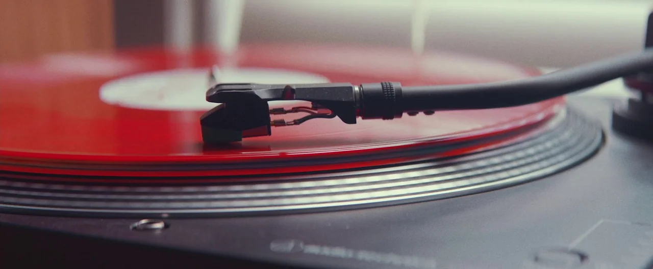 Poser (2021) movie still: closeup — A close-up of a red vinyl record spinning on a turntable platter. The tonearm is positioned at the edge of the…
