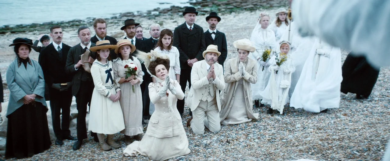 Slack Bay (2016) movie still: wide — A crowd of people in formal early 1900s clothing gathers on a pebble shoreline. Two individuals kneel in prayer…