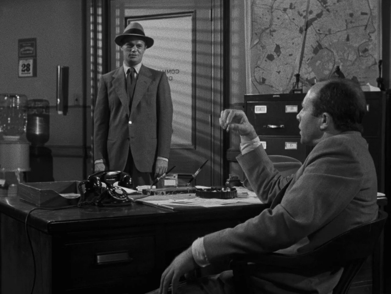 Pickup on South Street (1953) movie still: medium — An office from the 1940s features a wooden desk, a map on the wall, and venetian blinds. A man…