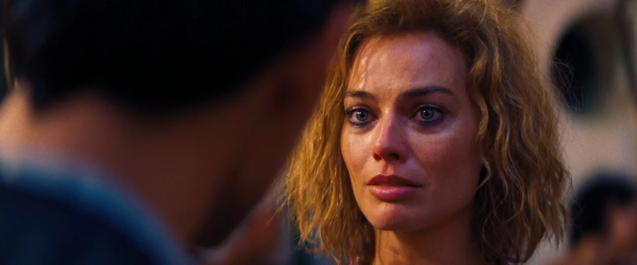 Babylon (2022) movie still: closeup — A dimly lit interior space contains a woman facing toward a person out of focus. The background shows hints of…