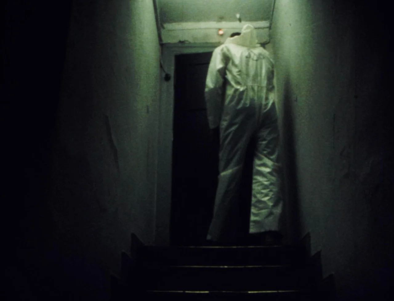 Nekromantik (1988) movie still: fullbody — A dark, narrow stairwell with visible concrete walls and a single closed door. A figure in white…