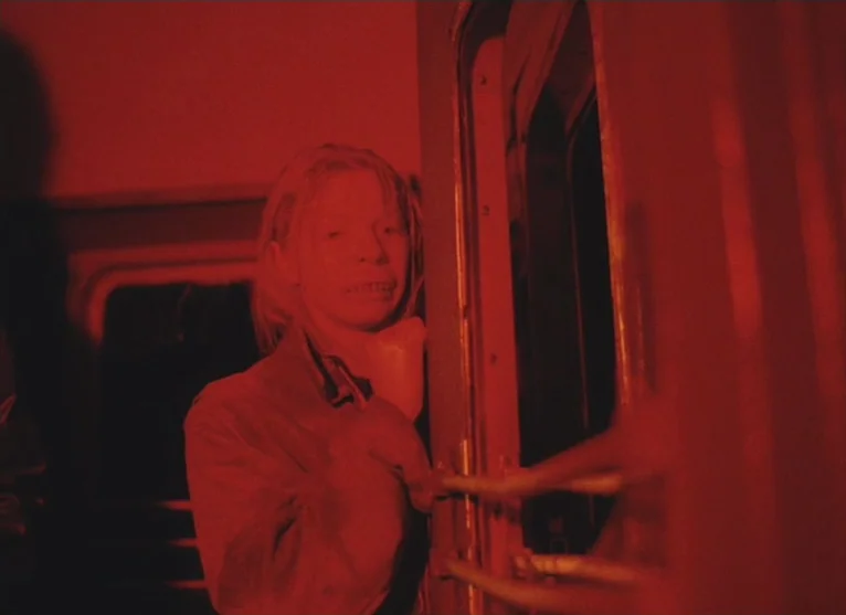 A Visitor to a Museum (1989) movie still: medium — A cramped train car interior is submerged in a saturated red light. A woman stands near the door…