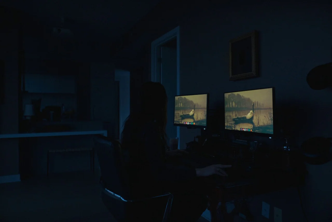 Red Rooms (2023) movie still: medium — A dark home workspace at night with a person seated before glowing monitors. The screens display a landscape…