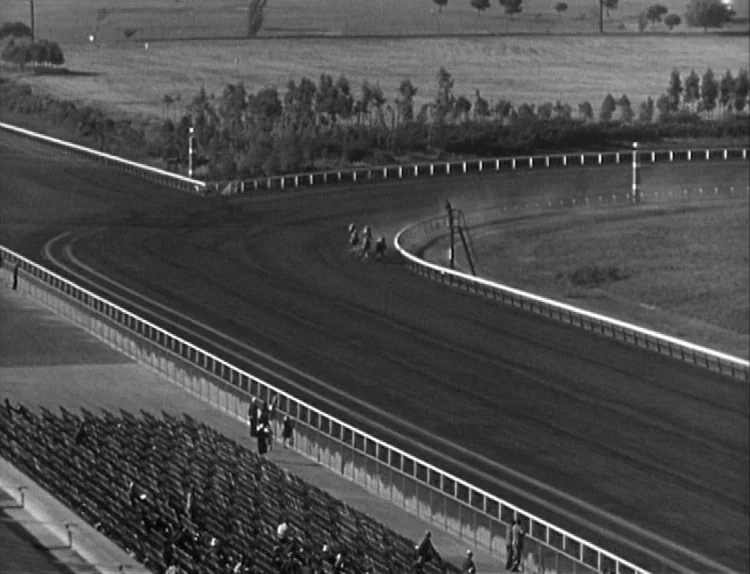 The Lady Eve (1941) movie still: establishing — A high-angle view of a large horse racing track with empty stadium seating. Several horses are…