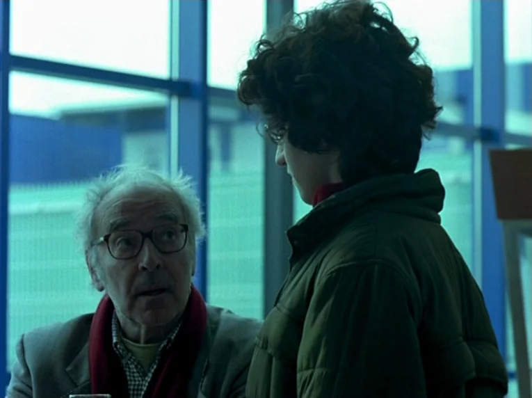 Notre Musique (2004) movie still: medium — A bright indoor space with large floor-to-ceiling windows framed by blue metal mullions. An older man…