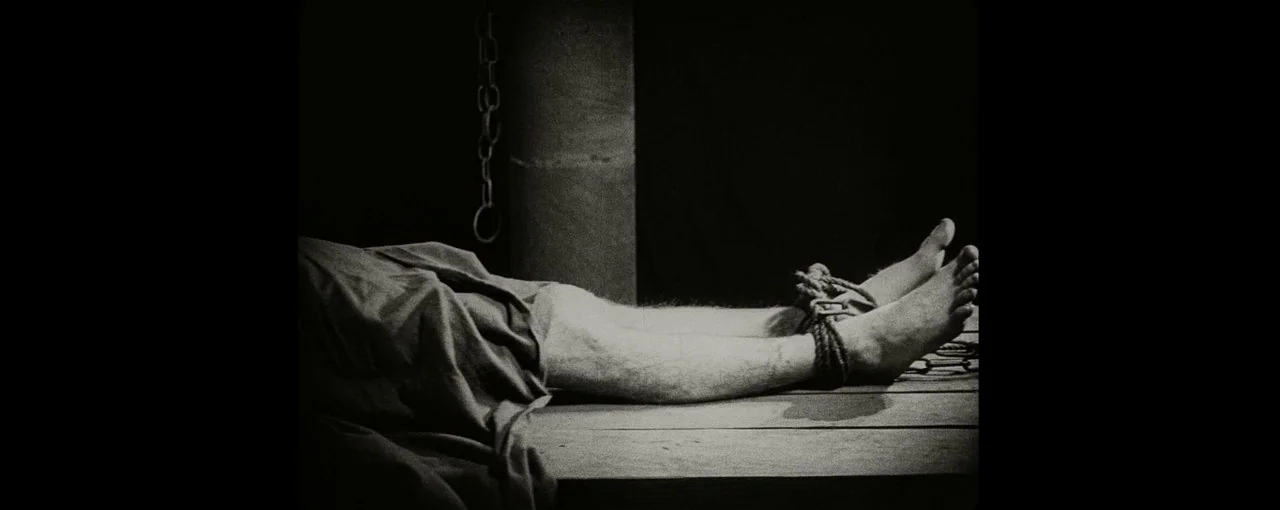 Lux Æterna (2019) movie still: closeup — A dark, spartan environment featuring a wooden platform and a hanging metal chain. A pair of male legs…