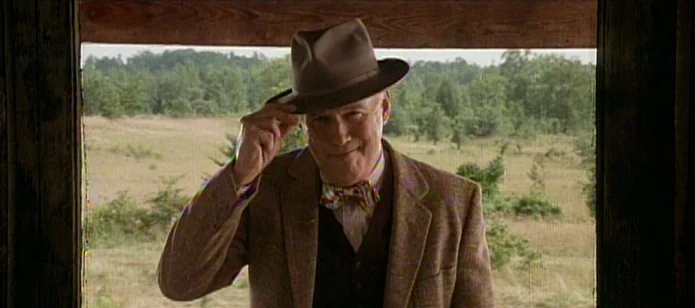 The Neon Bible (1995) movie still: medium — A man stands in a wooden frame looking out over an open grassy field. He tips his fedora hat with his…